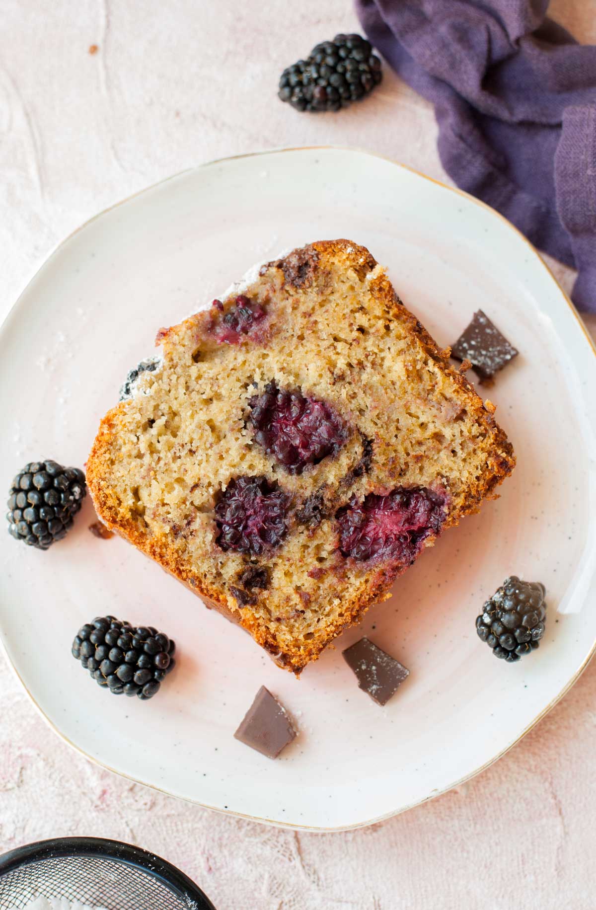 A slice of blackberry banana bread on a rosa plate, blackberries in the background.