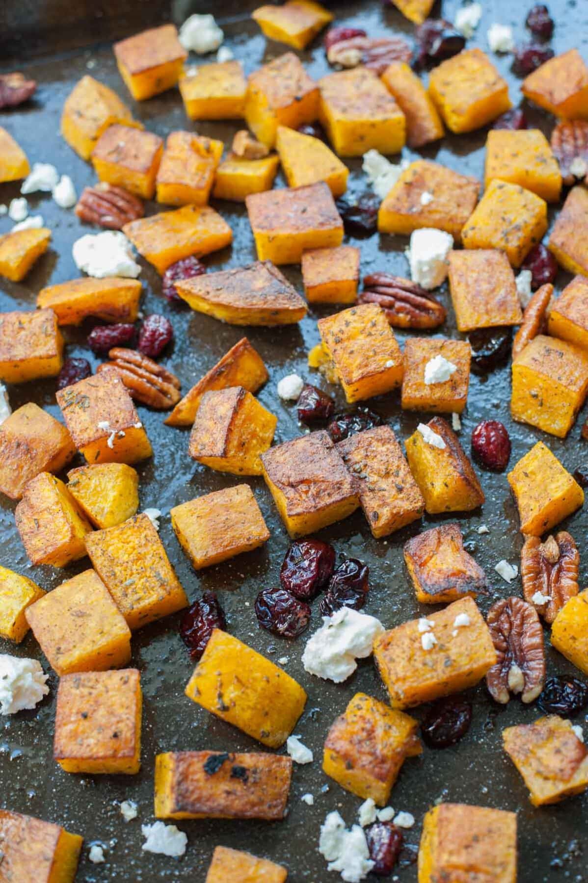 A close up picture of roasted butternut squash with cranberries, rosemary, pecans, and feta on a black baking tray.