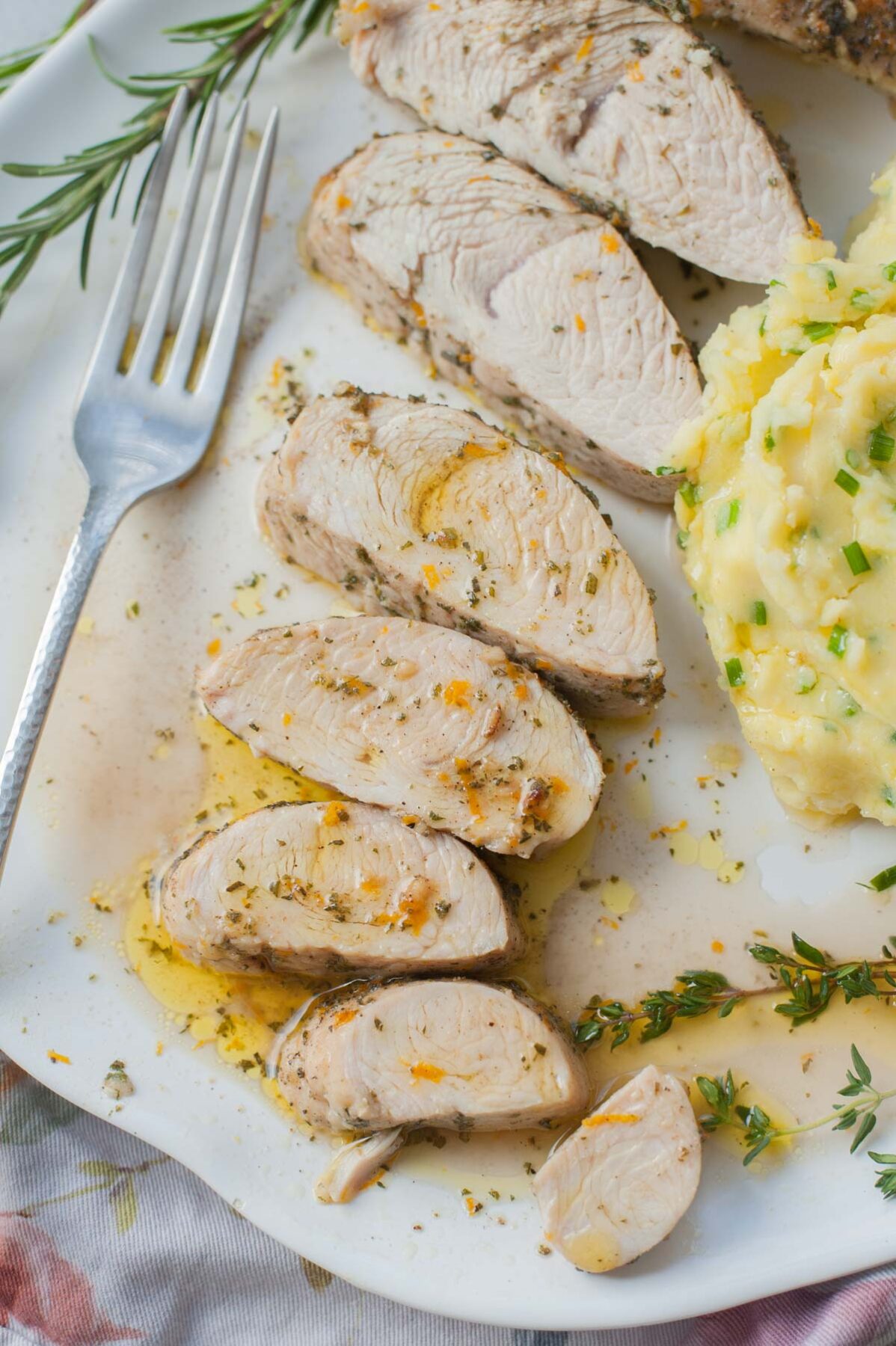Slices of baked turkey tenderloin on a white plate. Mashed potatoes and fresh herbs on the side.