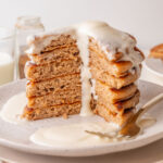 A tall stack of fluffy cinnamon pancakes with a generous cream cheese glaze dripping down the sides, served on a white speckled plate with a fork and extra glaze on the side. A large wedge is cut out, showing the soft, airy texture inside.
