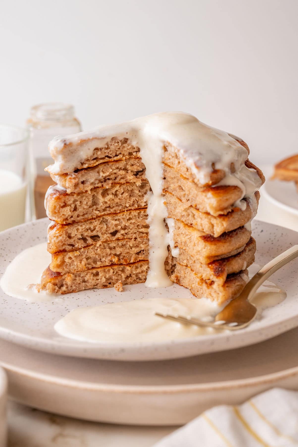 A tall stack of fluffy cinnamon pancakes with a generous cream cheese glaze dripping down the sides, served on a white speckled plate with a fork and extra glaze on the side. A large wedge is cut out, showing the soft, airy texture inside.