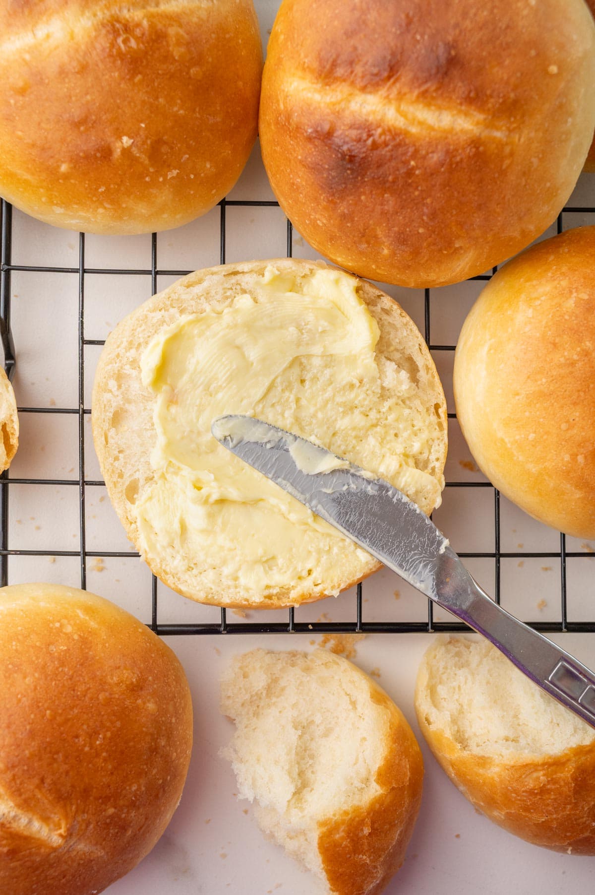 A freshly baked Brötchen sliced open and spread with butter, resting on a cooling rack. Surrounding it are more golden-brown rolls, with one partially torn to show the soft, fluffy inside.