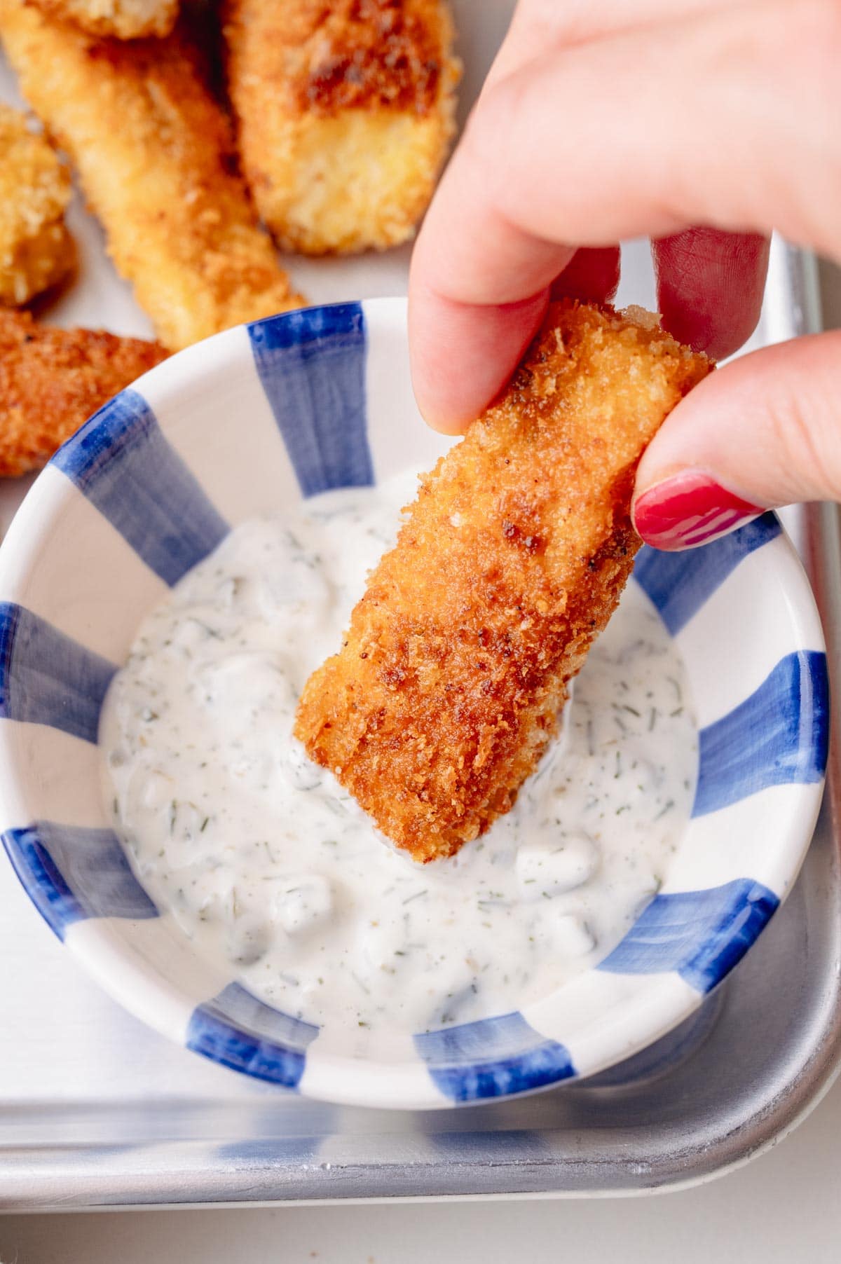 Hand dipping a crispy homemade fish stick into tartar sauce in a striped bowl.