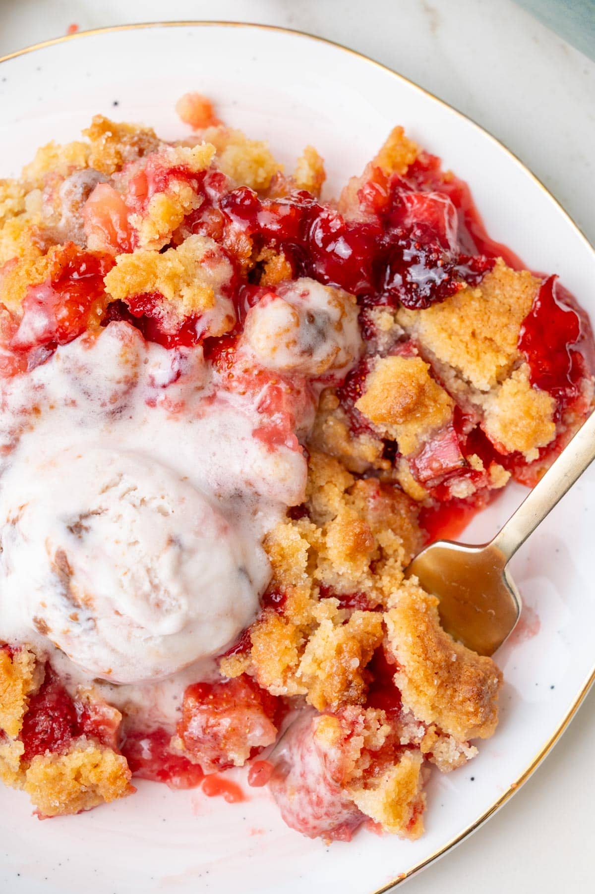Close-up of a serving of strawberry rhubarb crumble on a plate with melted ice cream and a spoon.