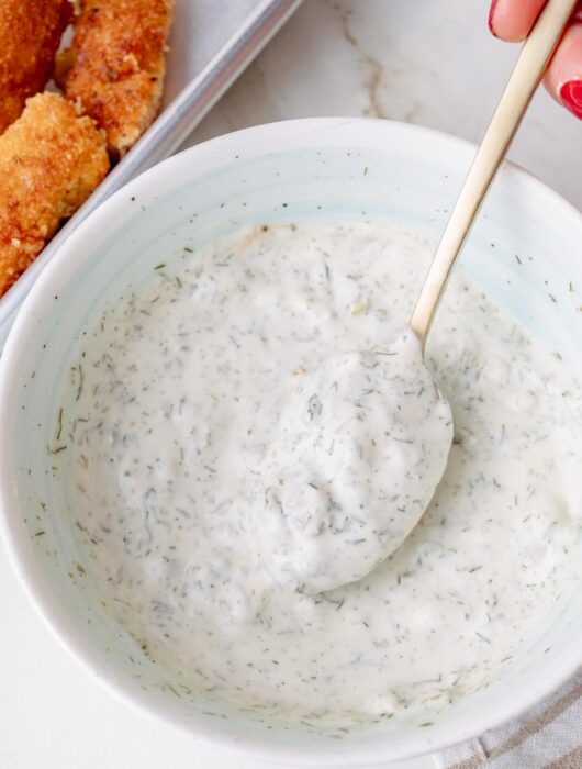 Close-up of a creamy homemade tartar sauce in a white bowl, with a spoonful lifted above the sauce.