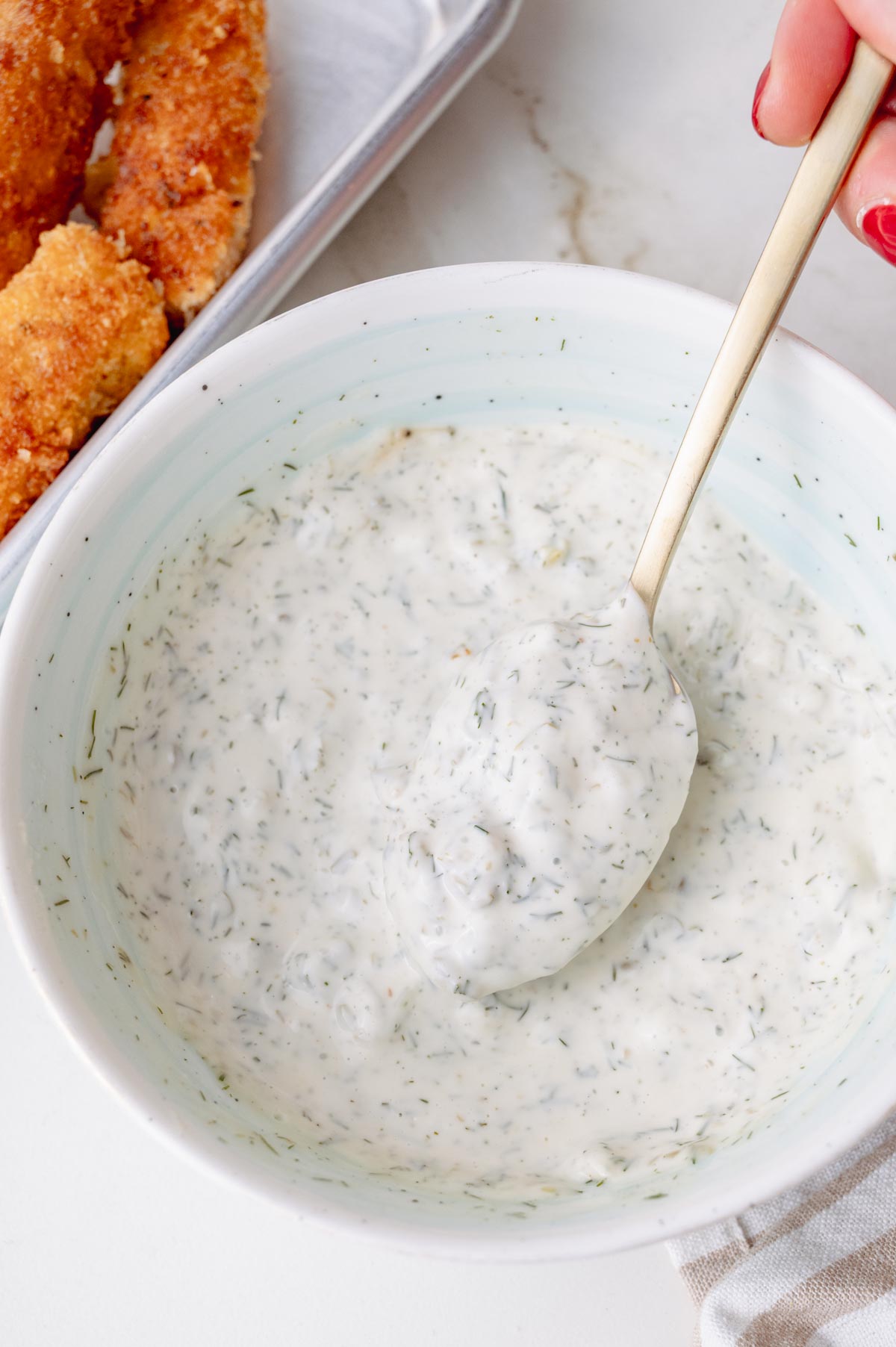 Close-up of a creamy homemade tartar sauce in a white bowl, with a spoonful lifted above the sauce. In the background, golden-brown fish sticks are visible on a metal tray.
