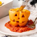 Stuffed pepper with carved face served on tomato sauce with knife cutting into the top.