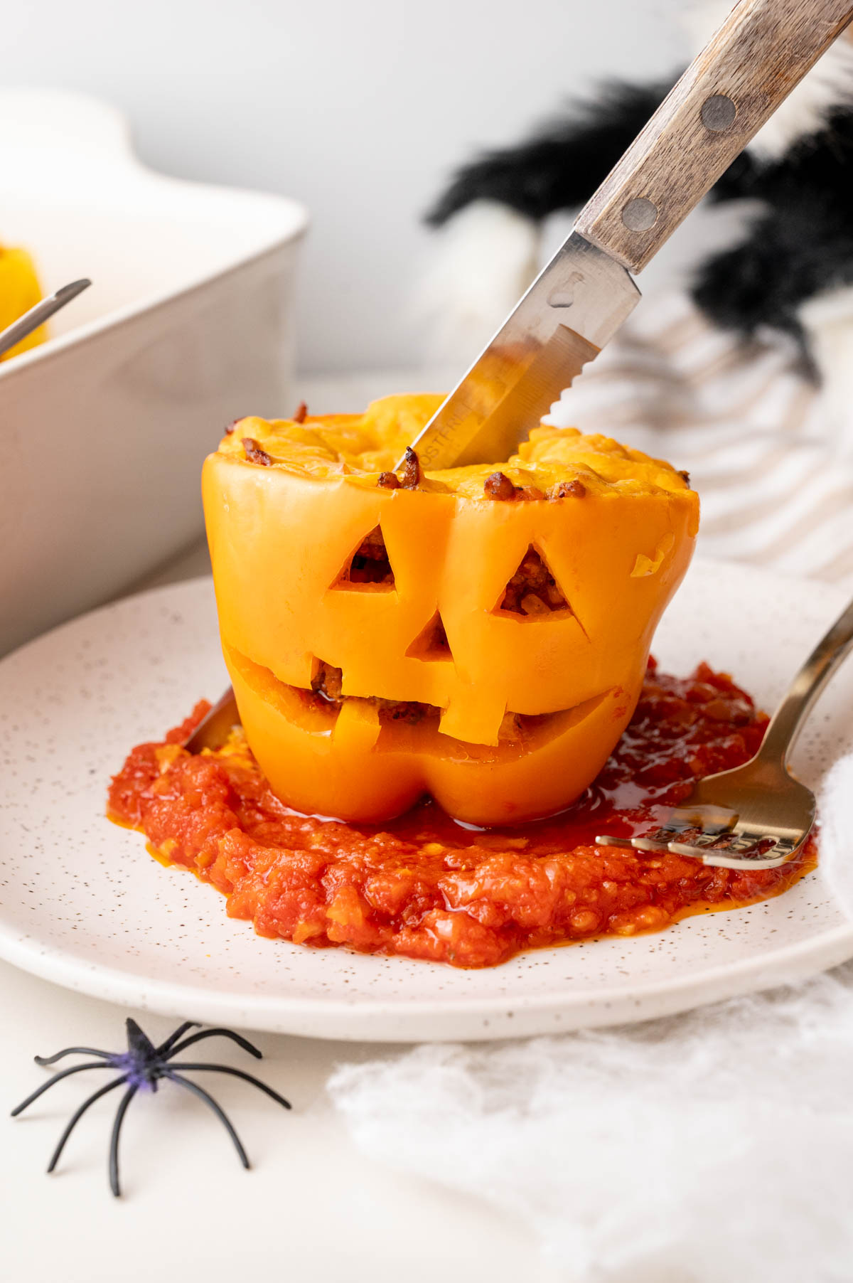 Stuffed pepper with carved face served on tomato sauce with knife cutting into the top.