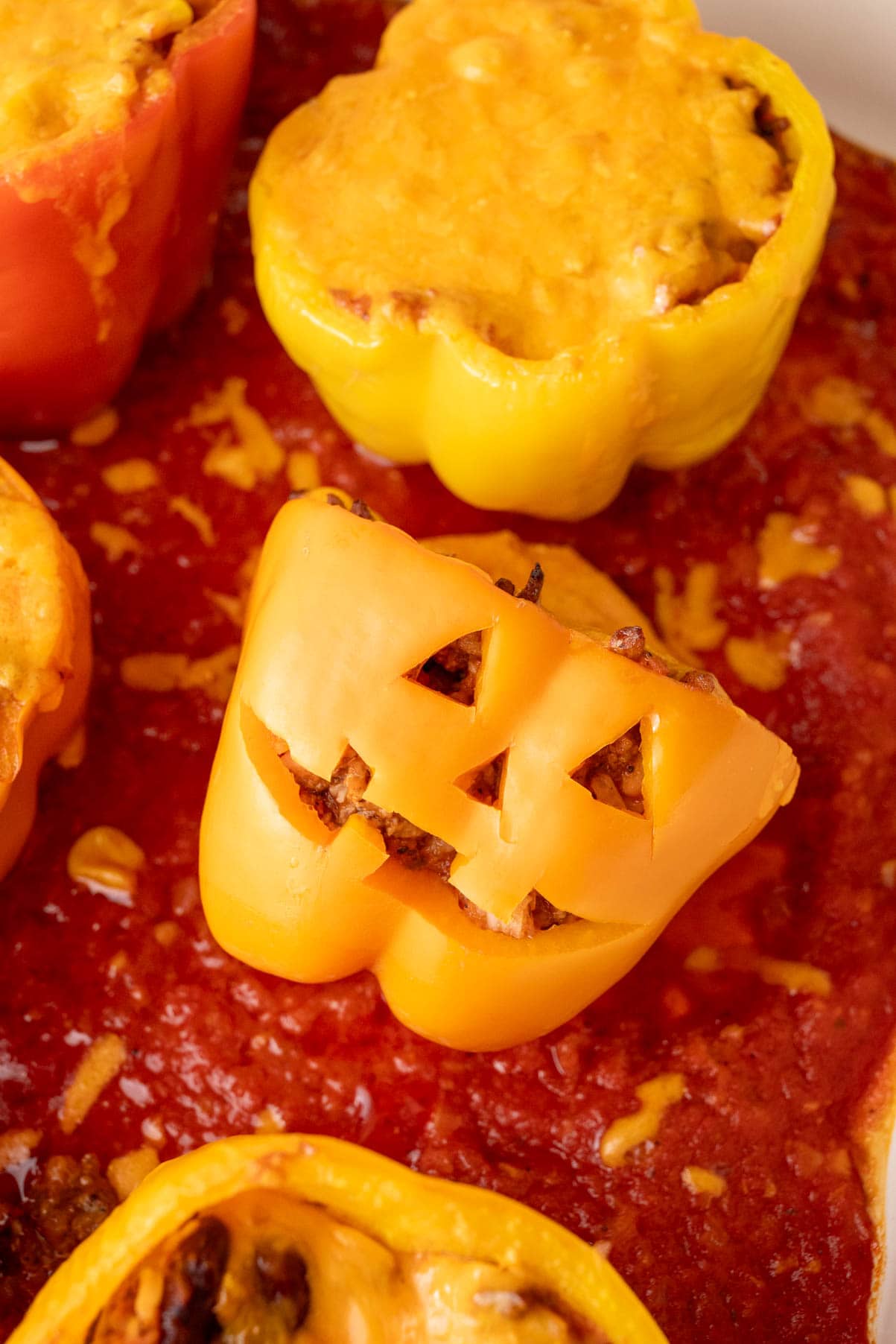 Baked jack-o'-lantern peppers lying on its side in a baking dish with tomato sauce showing carved face and melted cheese on top.