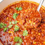 Hearty chili con carne with ground beef, beans, corn, and tomatoes simmering in a white Dutch oven, garnished with fresh cilantro and served with tortilla chips.
