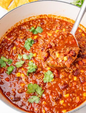 Hearty chili con carne with ground beef, beans, corn, and tomatoes simmering in a white Dutch oven, garnished with fresh cilantro and served with tortilla chips.