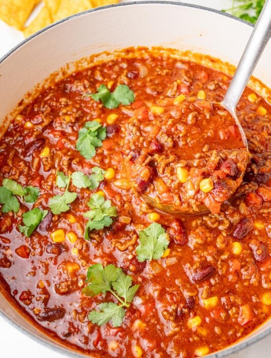 Hearty chili con carne with ground beef, beans, corn, and tomatoes simmering in a white Dutch oven, garnished with fresh cilantro and served with tortilla chips.