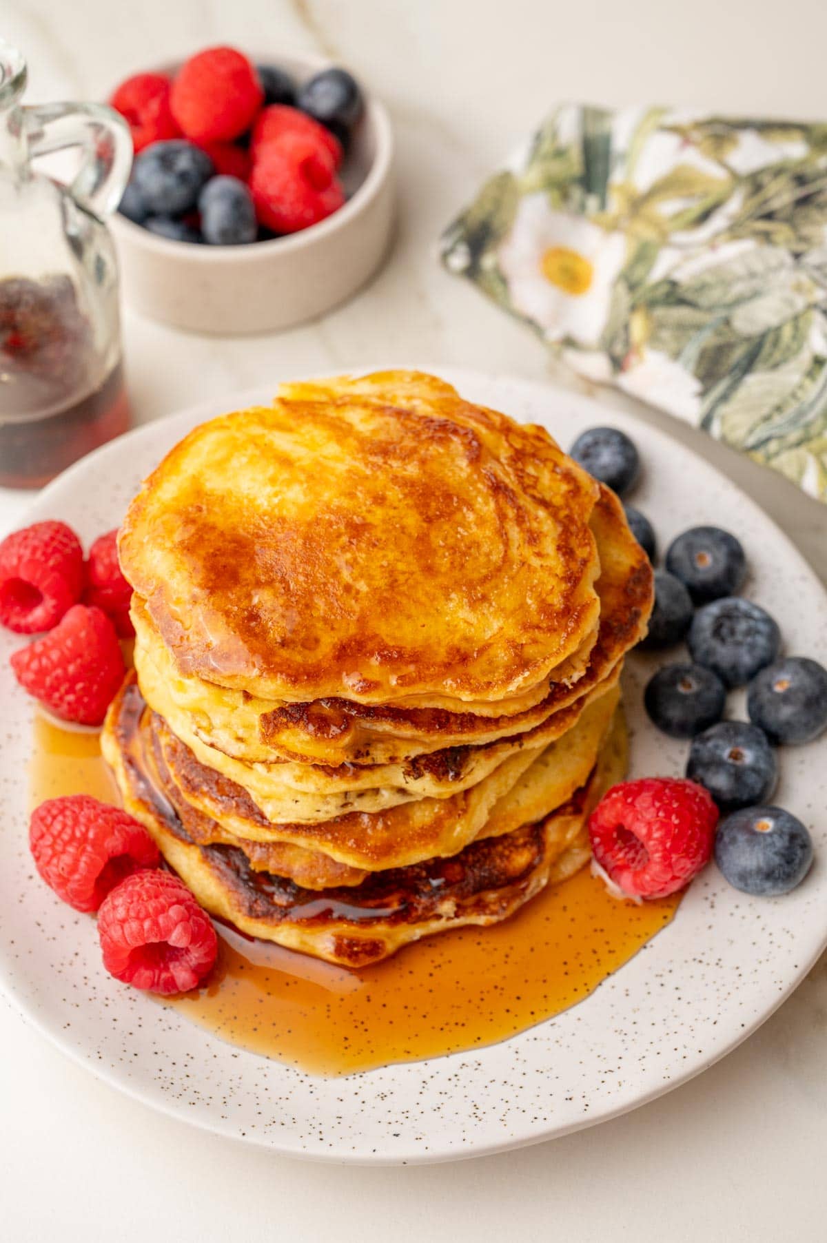 Stack of fluffy yogurt pancakes topped with maple syrup, served with fresh raspberries and blueberries.
