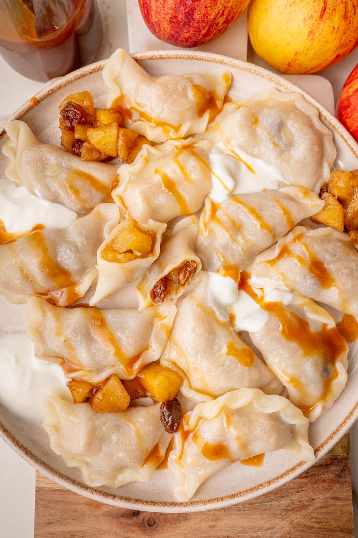 An overhead photo of apple pierogi on a beige plate served with caramel sauce and sour cream.