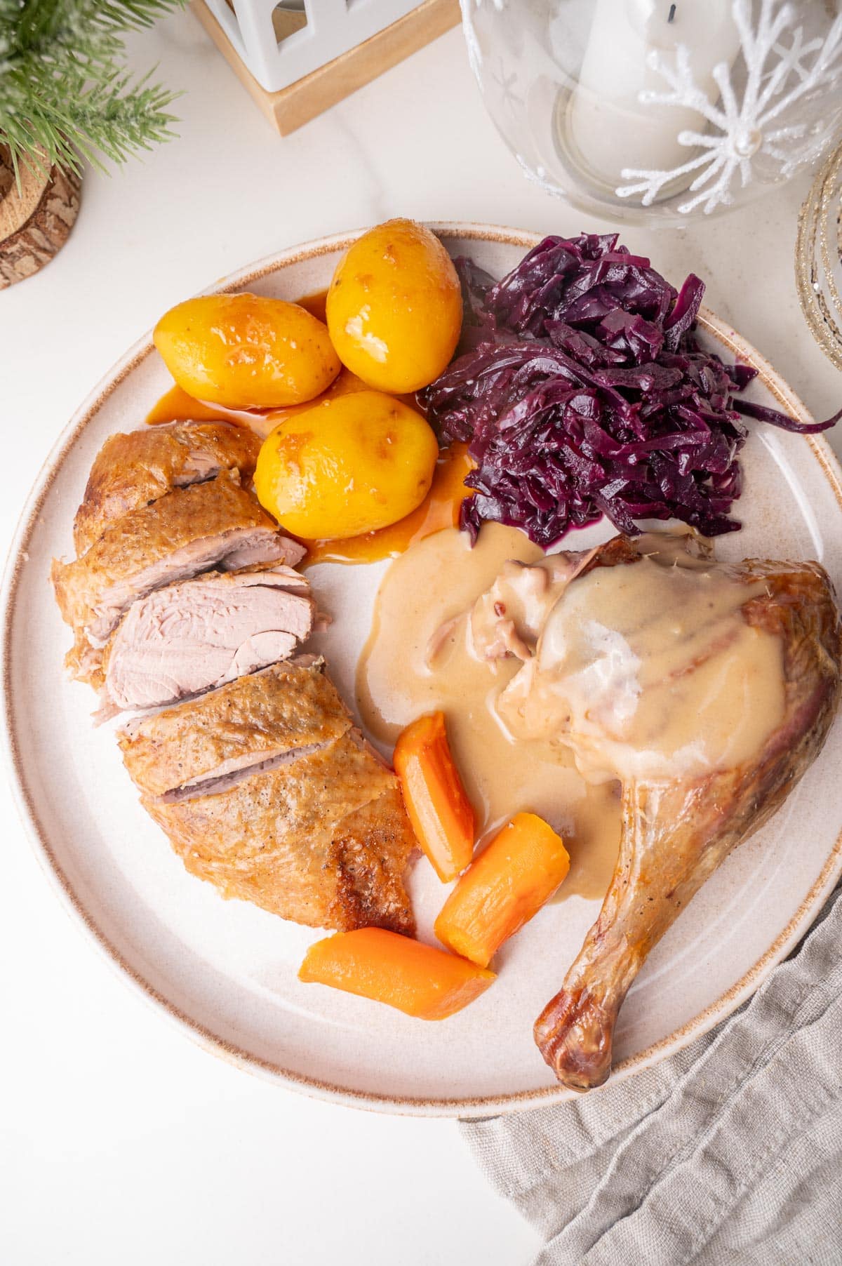 A duck breas and leg with gravy, caramel potatoes, red cabbage, and carrots on a beige plate.