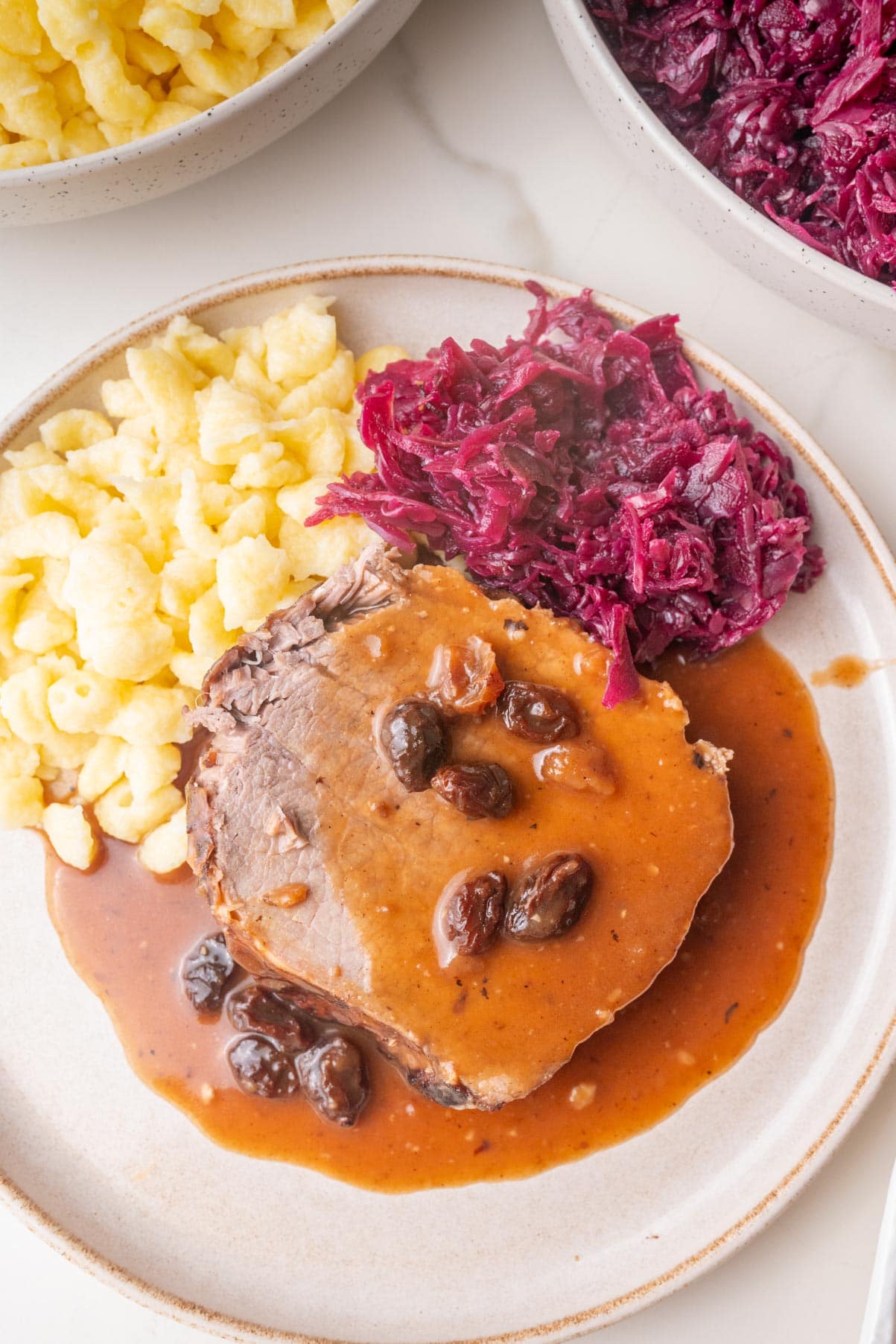 A slice of Sauerbraten with sauce and raisins, on a plate with Spätzle and red cabbage.